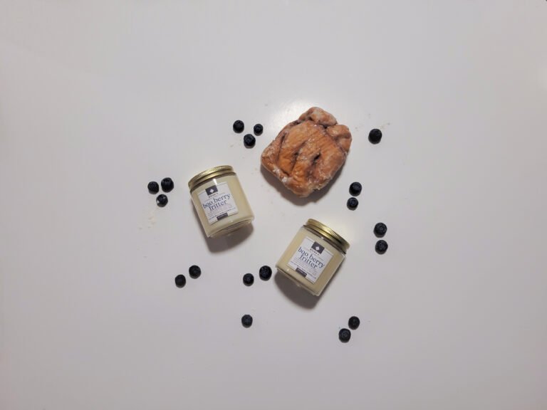 A glowing gold jar candle labeled "Boo Berry Fritter," surrounded by fresh blueberries, a cinnamon stick, a sugar-dusted fritter, and autumn leaves, all softly lit by the flicker of a crackling wick.