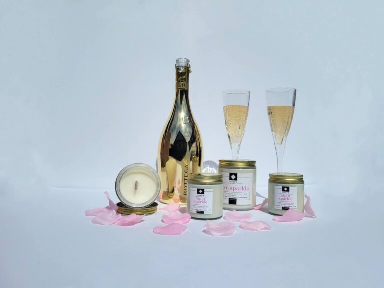 A collection of Alena’s Sip 'n Sparkle soy wax candles with gold lids and crackling wooden wicks. The elegant candles feature a soft glow, exuding hints of rose and sparkling notes, perfect for a luxurious ambiance.