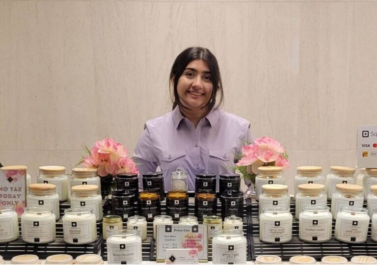 Alena standing proudly at her beautifully decorated booth, surrounded by gold jar candles, seasonal displays, and warm lighting, showcasing her Canadian Candle Company's hand-poured soy wax creations.