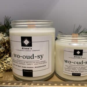 Alenas Wo- Oudsy Gold Lidded glass Jar Candles on a gold tray A rustic wooden candle jar labeled "Wo-oud-sy," surrounded by pieces of Brazilian rosewood, golden amber stones, and earthy foliage, evoking the warmth and richness of a summer bonfire.