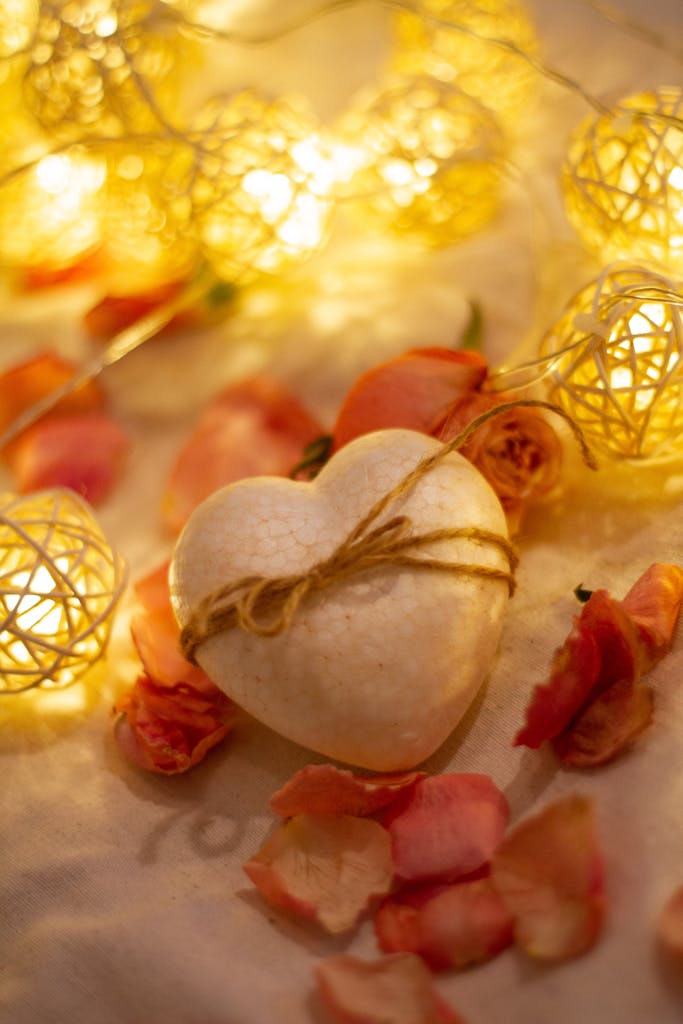 A heart-shaped stone wrapped in string amidst glowing fairy lights and rose petals.