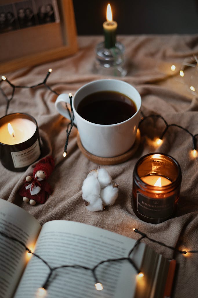 Warm and inviting still life setting with candles, coffee, and an open book illuminated by string lights.