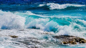 Vibrant blue ocean waves crashing against rocky shoreline under clear summer skies.